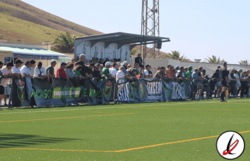 Jugadores y espectadores en la grada de un campo de fútbol en Lanzarote, con montañas y palmeras al fondo, durante un partido o evento deportivo.