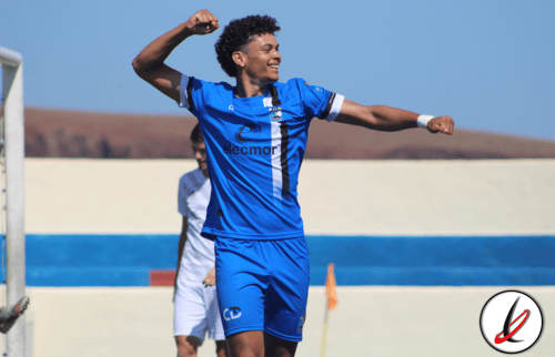 Jugador de fútbol en celebración en el campo, vestido con uniforme azul, en un estadio al aire libre con fondo de cielo azul y gradas.