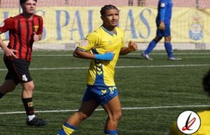 Jugador de fútbol en acción durante un partido en el campo de Lanzarote, vistiendo uniforme amarillo y azul, con otros jugadores y público en el fondo.