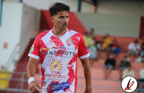 Jugador de fútbol en acción en el campo, vistiendo camiseta blanca y roja con patrocinadores, en estadio con espectadores en el fondo.