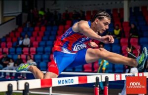Saltador en una pista de atletismo durante una competición, en pleno salto sobre la valla con estadio y espectadores en el fondo.
