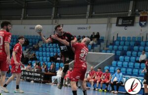 Balonmano en acción durante un partido, jugadores en movimiento en la pista cubierta del estadio, con público y banquillos en el fondo, en un encuentro deportivo en Lanzarote.