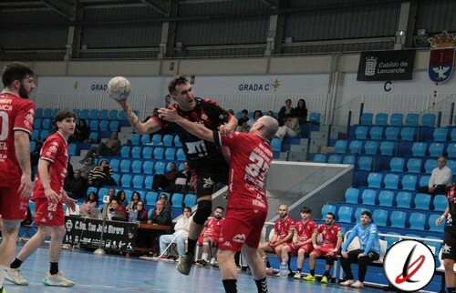 Balonmano en acción durante un partido, jugadores en movimiento en la pista cubierta del estadio, con público y banquillos en el fondo, en un encuentro deportivo en Lanzarote.
