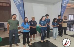 Grupo de jóvenes en una ceremonia, algunos con camisetas azules y otros con camisetas negras, en un espacio interior con banderas azules y un logo en la esquina inferior derecha.