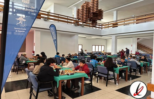 Sala de juego de ajedrez con varias mesas y jugadores jóvenes y adultos en un espacio interior bien iluminado y moderno.