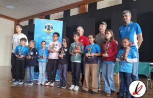 Grupo de niños y adultos en una ceremonia de premiación en un evento deportivo en un salón cubierto.