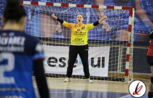 Guardameta en acción durante un partido de balonmano, con los brazos extendidos frente a la portería en un pabellón deportivo.