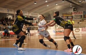 Jugadoras de voleibol en acción en un partido en interiores, con una jugadora en el centro intentando bloquear o recibir el balón mientras las otras dos se preparan para la jugada.