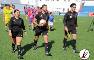 Árbitros de fútbol caminando en el campo de juego durante un partido en Lanzarote, con jugadores y espectadores en el fondo.