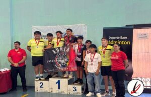 Grupo de jóvenes en el podio de un torneo de bádminton, con medallas, en una instalación deportiva cubierta, con banners y personal de organización presentes.