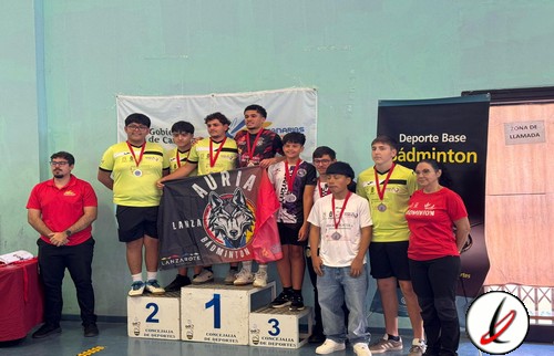 Grupo de jóvenes en el podio de un torneo de bádminton, con medallas, en una instalación deportiva cubierta, con banners y personal de organización presentes.