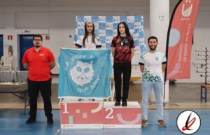 Cuatro deportistas en el podio de una competición de tiro con arco, con una bandera de "OB GRAN-K TOP SPIN" en el centro, en un evento indoor en Lanzarote.
