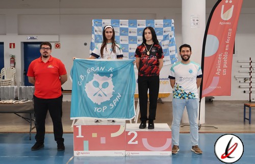Cuatro deportistas en el podio de una competición de tiro con arco, con una bandera de "OB GRAN-K TOP SPIN" en el centro, en un evento indoor en Lanzarote.