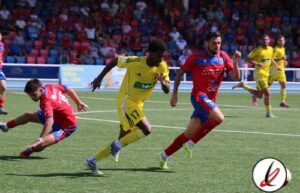 Jugadores en acción durante un partido de fútbol en un estadio con espectadores en las gradas.