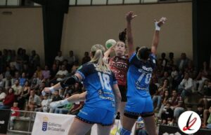 Jugadoras de balonmano en acción durante un partido en un pabellón con espectadores en las gradas.