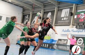 1. Jugadora de balonmano en acción durante un partido en un pabellón cubierto, vistiendo uniforme negro y rojo, con otras jugadoras y árbitros en el fondo.