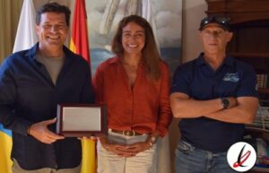 Tres personas sonriendo en una oficina, una de ellas sostiene un premio o reconocimiento en sus manos.