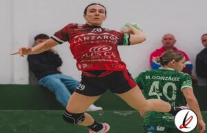 Jugadora de balonmano en acción, lanzando el balón en un partido en interiores, con espectadores en el fondo y otra jugadora en uniforme verde detrás.