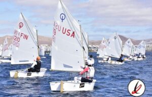 Nautas en regata de vela con varias embarcaciones en el agua, banderas de Estados Unidos y Finlandia en las velas, fondo de montañas y cielo parcialmente nublado.