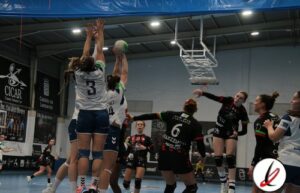 Balón en juego en un partido de voleibol femenino en una cancha cubierta, con jugadoras en acción, algunas saltando para bloquear y otras preparadas para recibir.