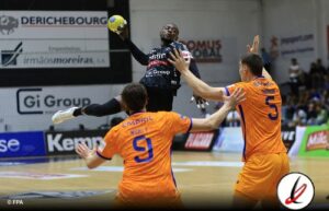 Balonmano en acción: jugador en camiseta negra saltando para lanzar el balón mientras dos defensores en camiseta naranja intentan bloquear su tiro en un partido en un pabellón cubierto.