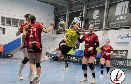 Atletismo femenino en acción durante un partido de balonmano en una cancha cubierta. La portera en uniforme amarillo intenta bloquear un disparo, mientras jugadoras con camisetas rojas y números 21, 4.