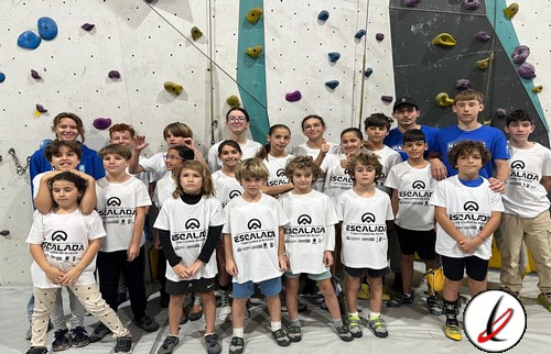 Escalada infantil en gimnasio de roca, grupo de niños y jóvenes con monitores, todos con camisetas blancas, en interior con paredes de escalada y agarres de colores.