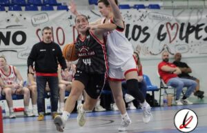 Jugadora de baloncesto en acción durante un partido en un gimnasio. La jugadora con camiseta negra y número 44 intenta anotar mientras una oponente en camiseta blanca la defiende. En el fondo, otros j.