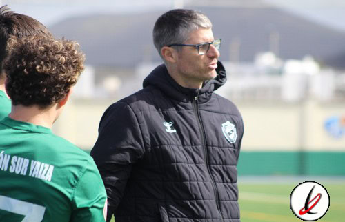 Entrenador de fútbol con gafas y chaqueta negra en el campo de entrenamiento, con jugador en camiseta verde en primer plano, en Lanzarote Deportiva.