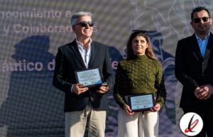 Premio de reconocimiento en un evento, con tres personas en el escenario, dos de ellas sosteniendo placas y una con gafas de sol. Fondo con texto y logotipo, en un ambiente formal y de celebración.