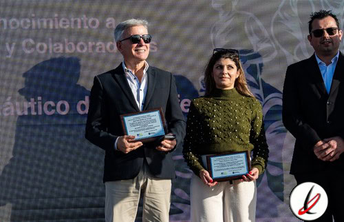 Premio de reconocimiento en un evento, con tres personas en el escenario, dos de ellas sosteniendo placas y una con gafas de sol. Fondo con texto y logotipo, en un ambiente formal y de celebración.