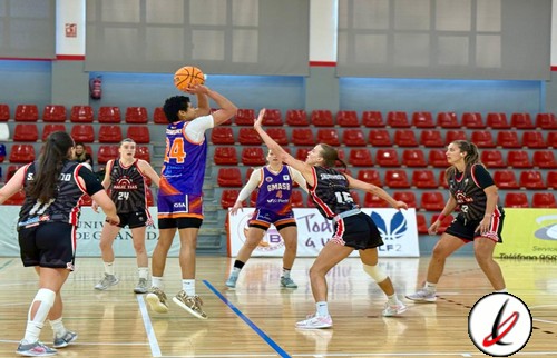 Balón de baloncesto en juego durante un partido femenino en una pista cubierta, con jugadoras en acción, público en el fondo y asientos rojos en la grada.