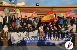 Bandera de España y banderas de otros países, grupo de jóvenes en un evento deportivo, algunos con camisetas negras, en un entorno al aire libre con fondo de estructura de madera y cielo despejado.