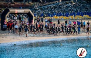 Personas en la playa participando en un evento deportivo con un arco de salida y sombrillas azules y amarillas en Lanzarote.