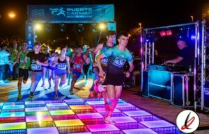 Correr en una carrera nocturna en Lanzarote, con participantes jóvenes en ropa deportiva, cruzando una pista de luces LED en un evento organizado en un entorno iluminado y festivo.