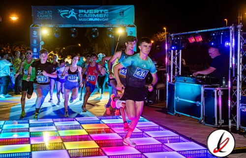 Correr en una carrera nocturna en Lanzarote, con participantes jóvenes en ropa deportiva, cruzando una pista de luces LED en un evento organizado en un entorno iluminado y festivo.