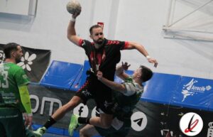 Balonmano en acción: jugador en el aire lanzando el balón con fuerza durante un partido en una cancha cubierta. Jugadores rivales intentan bloquear el tiro en un entorno deportivo con anuncios publici.