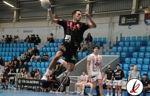 Balonmano en acción en un partido en Lanzarote, jugador saltando para lanzar el balón, con espectadores y banquillos en el fondo en un pabellón deportivo.
