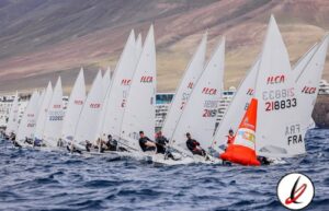 Navegantes en regata de vela con varias embarcaciones en el agua, montados en sus barcos con velas blancas y un inflable naranja en el extremo. Montañas y cielo despejado en el fondo.