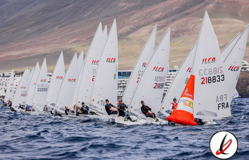 Navegantes en regata de vela con varias embarcaciones en el agua, montados en sus barcos con velas blancas y un inflable naranja en el extremo. Montañas y cielo despejado en el fondo.