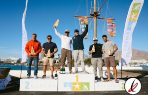 1. Hombres en el podio de una regata, con el barco y banderas de fondo, celebrando su victoria en Lanzarote.