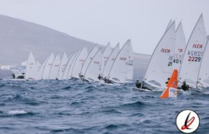 Navegantes en regata de vela en el mar con varias embarcaciones y velas blancas, en un día nublado y con olas moderadas.
