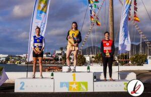 Atletas en el podio de una competición deportiva en Lanzarote, con banderas y un puerto de fondo.