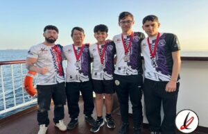 Grupo de cinco jóvenes deportistas con medallas en un barco, con vista al mar y cielo despejado al atardecer.