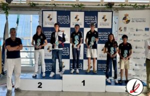 Atleta en podio de natación con medallas, en una ceremonia de premiación en un evento de la Sección Náutica de Tenerife, con fondo de banners y un entrenador a la izquierda.