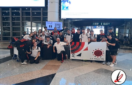 Jugadores de fútbol en grupo en aeropuerto con cartel del equipo y maletas, listos para viajar.