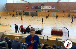 Pista de bádminton en un gimnasio cubierto con jugadores practicando y espectadores en las gradas. Niños y adultos participan en actividades deportivas, con mesas y premios en el fondo.