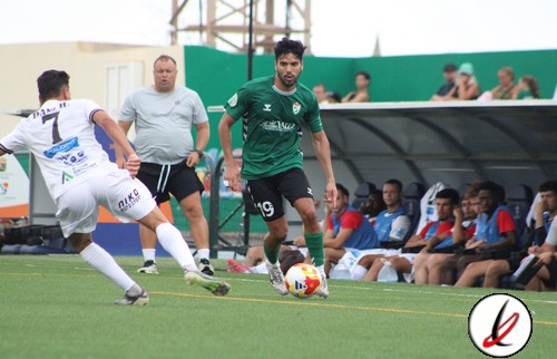 Jugador con camiseta verde controlando el balón en un partido de fútbol en Lanzarote, con jugadores rivales y banquillo en el fondo.