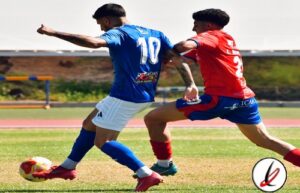 Jugadores de fútbol en acción durante un partido en un campo al aire libre, uno con camiseta azul y el otro con camiseta roja, luchando por el balón.