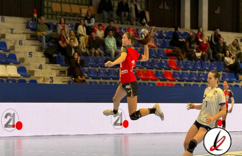 Saltadora en acción durante un partido de balonmano en un pabellón deportivo con espectadores en las gradas. La jugadora lleva un uniforme rojo y negro, y está en pleno salto con la pelota en la mano.
